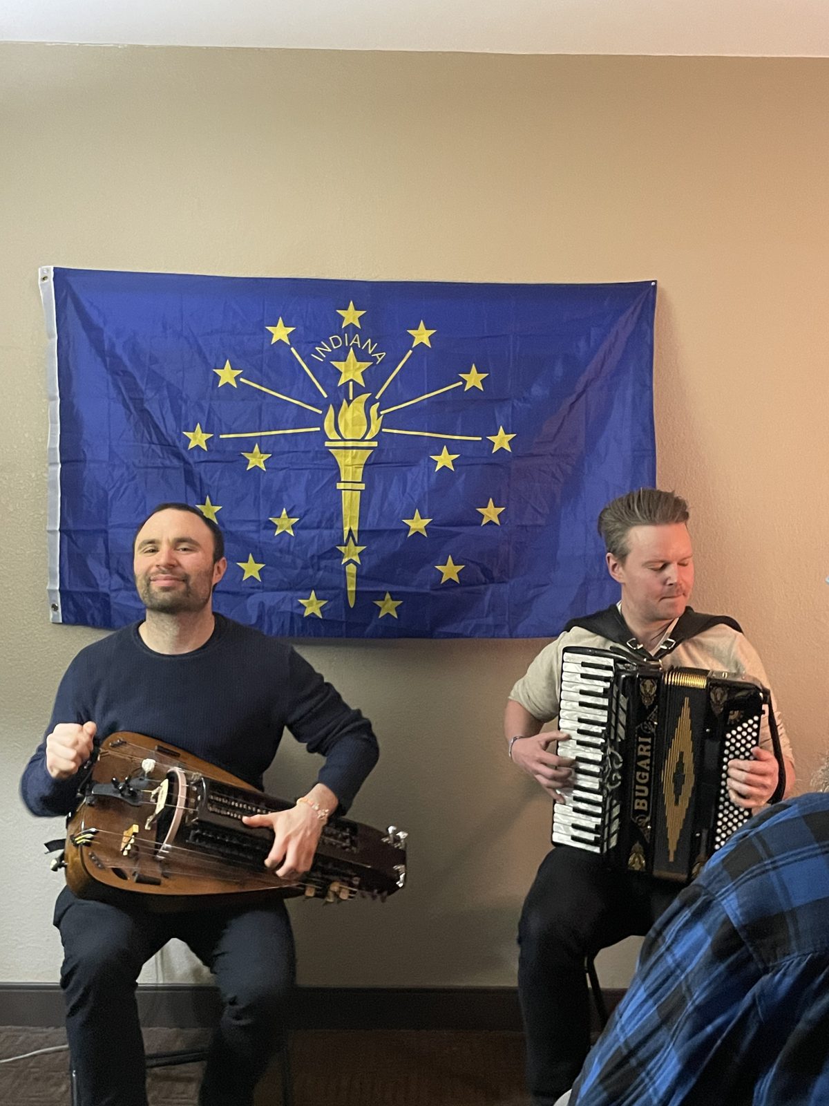 A duo on hurdy gurdy and accordion in front of the Indiana flag